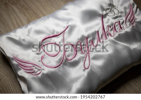 Machine embroidery on a white satin robe for the bride.
Inscription bride in Bulgarian and wings for decoration with pink thread. Close-up photo of a wooden background.
