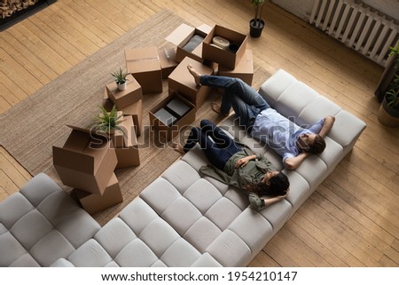 Top view happy couple tenants homeowners relaxing on couch on moving day in living room with cardboard boxes, man and woman resting after relocation in first own apartment, mortgage or rent Royalty-Free Stock Photo #1954210147