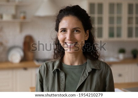 Head shot portrait smiling woman looking at camera, posing for profile picture, standing in modern kitchen at home, confident positive businesswoman making video call, involved in internet meeting