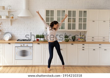Overjoyed young woman tenant dancing, jumping in modern kitchen at home, excited by relocation in first own apartment, happy attractive young female having fun, moving to favorite music alone Royalty-Free Stock Photo #1954210318