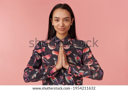 Young lady, pretty asian woman with dark long hair. Wearing black shirt with birds. Emotion concept. Keeps palms together in prayer, namaste. Watching at the camera isolated over pink background