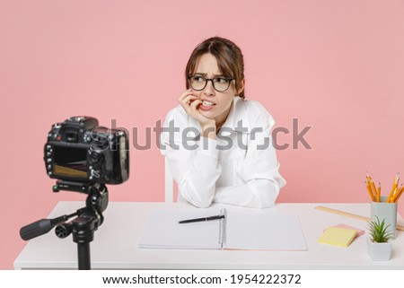 Worried puzzled sad woman tutor teacher in shirt glasses sit at desk gnawing nails conducting online lesson seminar recording video on camera isolated on pink background. Distance education concept
