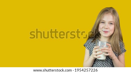 A cute little girl with blonde hair is holding and drinking milk from a glass. It stands on a yellow background. Milk is good for children. She is healthy and happy. Banner. Soft focus. Copy space.