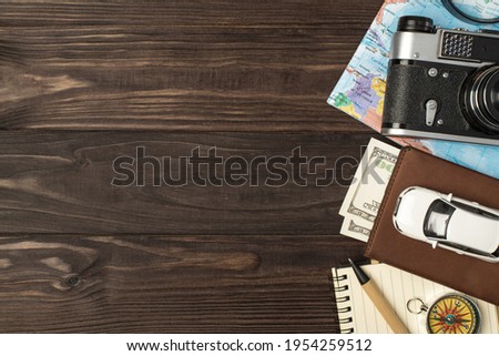 Top view photo of car model on passport cover with dollars camera map magnifier compass notebook and pen on isolated wooden table background with copyspace on the left