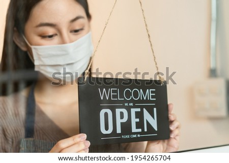 Smiling young asian owner retail,coffee shop woman turning sign board to open wearing face mask,protect to pandemic of coronavirus,reopen store after close lockdown quarantine in covid to new normal.