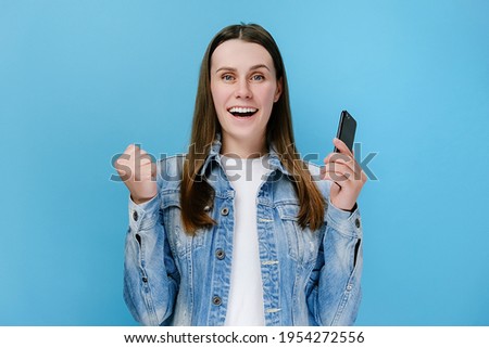 Excited cheerful young woman holds smartphone receive sms with great news feels happy make yes gesture, wears denim jacket, isolated on blue background. Moment of victory showing sincere emotions