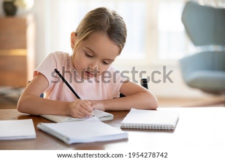 Smart small 7s Caucasian girl child sit at table at home write in exercise book prepare homework for school. Little kid handwrite in notebook do task assignment. Learning, education concept. Royalty-Free Stock Photo #1954278742