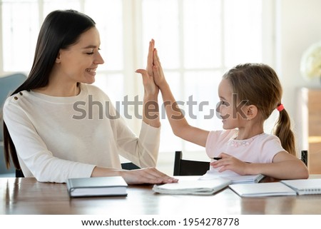 Smiling young Caucasian mother and little 7s daughter give high five for finished homework assignment. Happy loving mom and small girl child celebrate good study result. Education, learning concept. Royalty-Free Stock Photo #1954278778