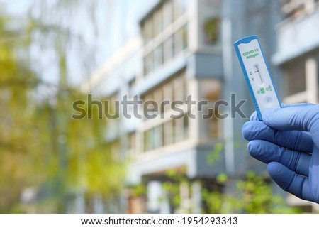 Hand holds quick test outdoors, with office, school or kindergarden building out of focus. Express COVID19 test, Schnelltest is rapid corona test in German. Selbsttest means self-test for coronavirus.