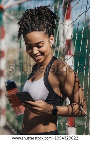 Body activity check, good results, app and social networks, sports blog. Smiling young african american woman in sportswear with headphones holds bottle of water typing on phone at stadium, vertical