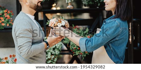 Attractive and smiling man client buying bouquet of white fresh flowers from market stall store. Millennial woman salesman in apron giving composition to customer near studio, panorama, cropped
