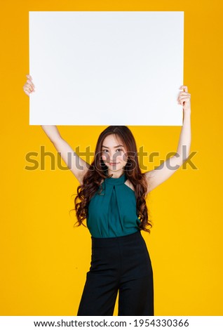 Cute and pretty curly hair Asian female brunette holding white  blank board over her head with a joyful for advertising and banner use purpose, studio shot isolated on bright yellow background