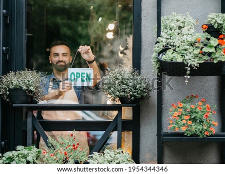 Good morning, start of working day, opening small business after covid-19 pandemic. Smiling millennial man in apron in flower shop with plants in pots and glass door turns sign is open, copy space