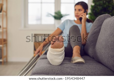 Young woman with broken leg in cast lying relaxing on sofa with crutches nearby and talking on phone at home, selective focus. Injury, trauma, recovery, rehabilitation concept