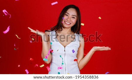 Funny young and cure Asian girl feels excited and cheerful while playing with flying confetti from the above position. Studio shot on a red background.