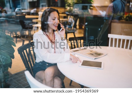 Side window view of happy Caucasian customer checking touch pad notification while talking in cafe interior, successful female blogger connecting to wifi internet during morning in coffee shop