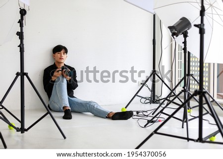 Asia young handsome successful male photographer wears black long sleeve shirt hold DSLR camera in hand while sit on floor leaning on white background checking photo from album in indoor photo studio.