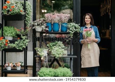 Woman florist reopening shop after covid-19 pandemic, delivery service for clients. Happy young female in apron holds bouquet of tulips near front door of store with vases and pots of diverse flowers Royalty-Free Stock Photo #1954379830