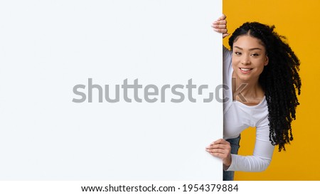 Positive Black Lady Peeking Out Of White Advertisement Board, Smiling Young African American Woman Holding Blank Billboard With Copy Space For Your Design Or Offer, Standing Over Yellow Background