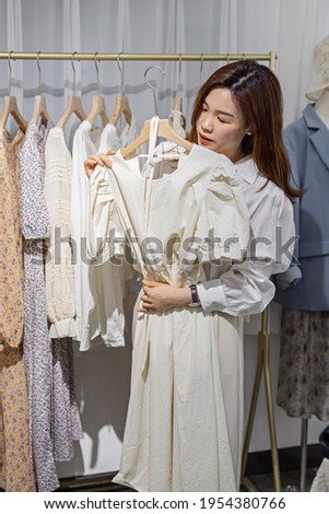 portrait of happy female asian entrepreneur working in her modern store with womenswear clothes, young chinese woman owner or consultant holding fashionable clothing while standing in brandy shop
