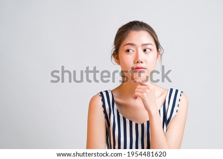 Portrait of young charming Asian woman looks pensive and curious thinking up and look sideways at grey background with copy space to the left, concept pensive thought woman, curious thinking woman. Royalty-Free Stock Photo #1954481620