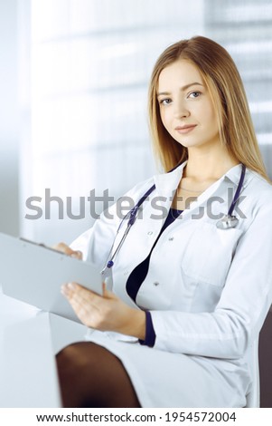 Young optimistic woman-doctor is using a clipboard, while sitting at the desk in her cabinet in a clinic. Portrait of friendly female physician woman with a stethoscope. Perfect medical service in a