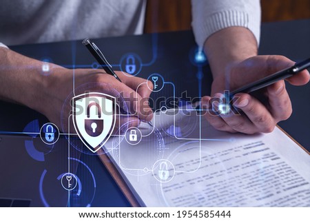 A businessman in casual wear signing the contract to prevent probability of risks in cyber security. Checking the phone. Padlock Hologram icons over the working desk.