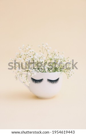 Creative white mug of flowers with long eyelashes on beige background.