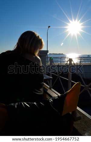 Photography of a woman sitting backwards in front of the river whit a big sun in the sky feflecting on the water