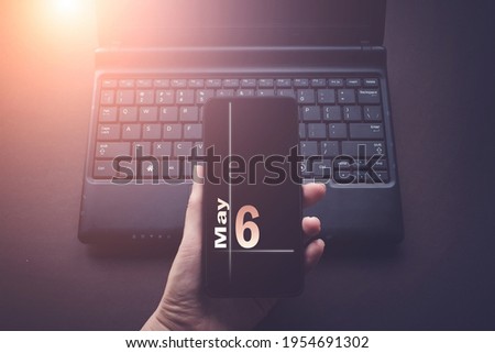 May 6th. Day 6 of month, Calendar date. Hand Holding Mobile Phone on Laptop Computer dark background with sunshine.  Spring month, day of the year concept