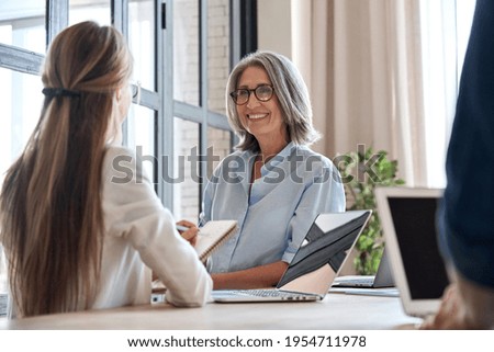 Happy mature middle 60s aged smiling lady boss manager teacher talking to assistant or student meeting in office space. Company team working together with the laptops and assistant writing notes. Royalty-Free Stock Photo #1954711978