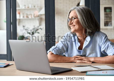 Senior adult mid 50s age watching at window at home sitting at table with laptop. Feeling happy and smiling to thoughts about future positive vision of successful training career after learning.