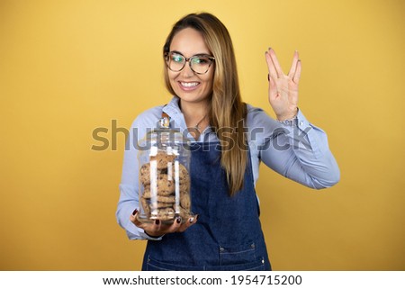 Young hispanic woman wearing baker uniform holding a cookies jar doing hand symbol
