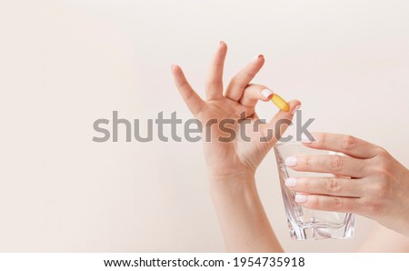 Feminine hands with trendy manicure holding glass of water and yellow capsule of vitamin d3 or fish oil omega. Neutral background with copy space. Wellbeing concept. Royalty-Free Stock Photo #1954735918
