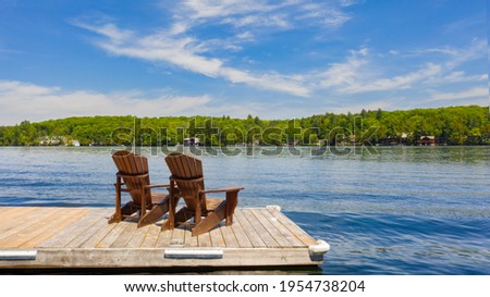 Two Adirondack chairs on a wooden dock facing a lake in Muskoka, Ontario Canada during a sunny summer morning. Cottages are nested between trees across the water. Royalty-Free Stock Photo #1954738204