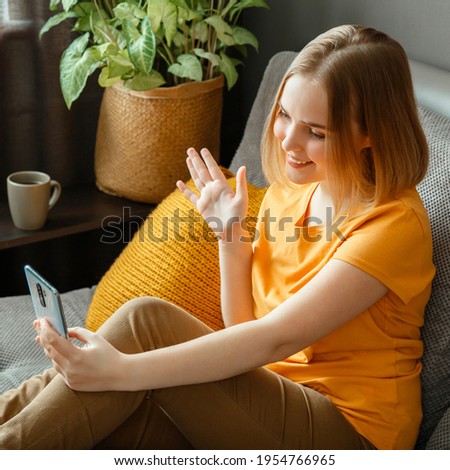 Happy young beautiful woman greets, wave by hand greeting, talking by video call using smartphone while sitting in living room. Teen girl make video call communication in home interior. Square crop
