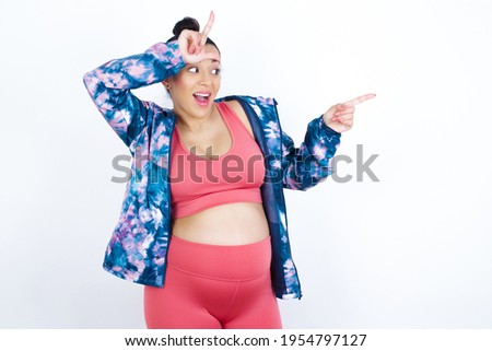 young beautiful Arab pregnant woman in sports clothes against white wall showing loser sign and pointing at empty space