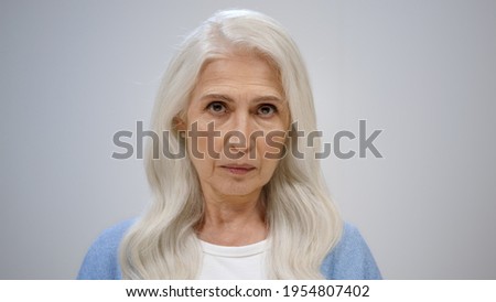 Focused senior woman looking camera inside. Serious old lady wagging finger on grey studio background. Portrait of disappointed female person gesticulating indoors.