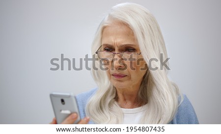Old woman reading news on mobile phone in studio. Focused aged lady fixing glasses indoors. Annoyed female person looking to screen of smartphone on grey background.