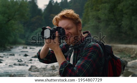 Smiling hiker shooting nature landscape in mountains. Young photographer using photo camera outdoor. Excited redhead man looking at photos on digital camera after shooting mountain river