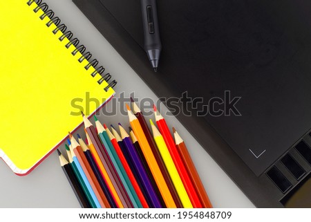 Digital stylus pen on digital graphic tablet for illustrators and designers opposite colored drawing pencils next to a wired notebook . Top view, free space for text