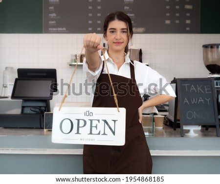 Happy barista standing at coffee shop counter proudly showing open sign board smiling. Beautiful woman in apron looking at camera welcome to her local small cafe business.   