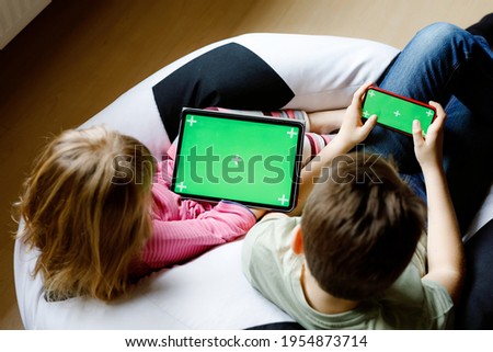 Kid boy and little girl gaming or using social media on mobile phone and tablet with chroma key background. Two children holding smartphone and laptop with green screen for commerce, new app.
