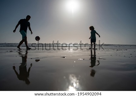 Father and son play soccer or football on the beach, silhouette on sunset. Dad and child having fun outdoors