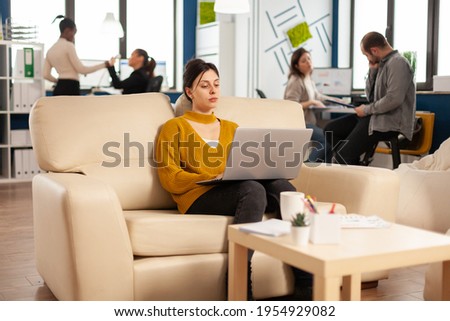 Succesfull business woman working on laptop on start up project sitting on cozy couch. Multiethnic professional company people coworkers discussing task in modern workplace.