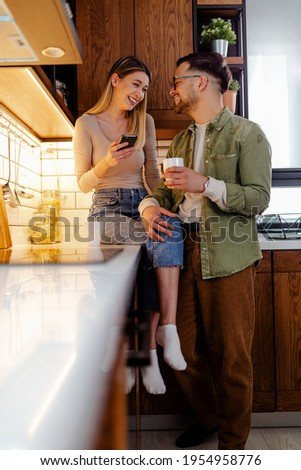 Young couple enjoying in cozy kitchen, woman holding smartphone sitting on worktop talking to her man, watching funny videos and drinking a coffee together.