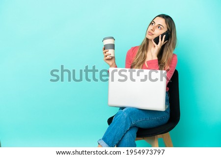 Young caucasian woman sitting on a chair with her pc isolated on blue background holding coffee to take away and a mobile