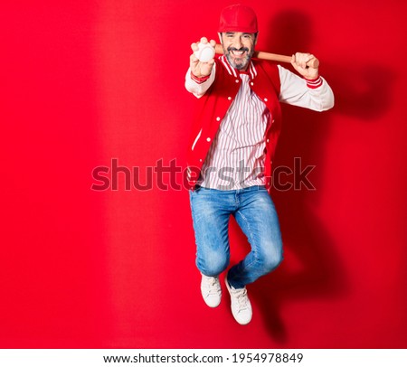 Middle age handsome man wearing sporty clothes smiling happy. Jumping with smile on face playing baseball using bat and ball over isolated red background