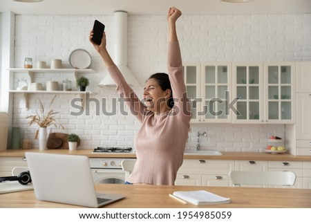 Excited latina woman freelancer raise hands up hold phone scream hooray in delight celebrate finish work on difficult project. Euphoric young lady remote employee meet deadline with all the work done Royalty-Free Stock Photo #1954985059