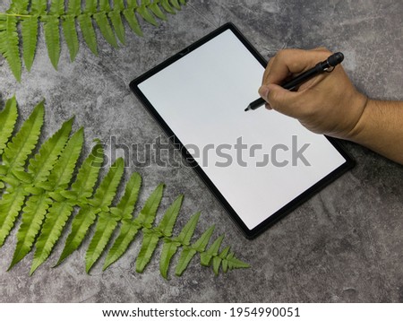 Hand holding pen writing tablet screen white Placed on a black cement floor Decorated with fern leaves